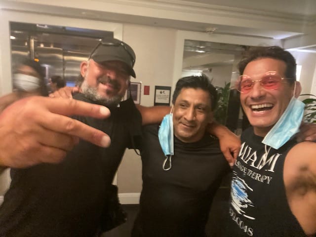 Three men posing for a photo indoors smiling and wearing casual attire Two have masks hanging around their necks and one is making a peace sign with his hand