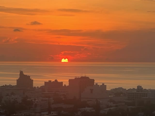 A sunset over a cityscape with orange and yellow hues in the sky buildings silhouetted in the foreground and a calm body of water in the background | Miami Massage Therapy South Beach Sunrise