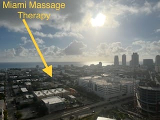 A city skyline view with Miami Massage Therapy labeled and an arrow pointing to a building in the foreground The sky is partly cloudy with the sun shining