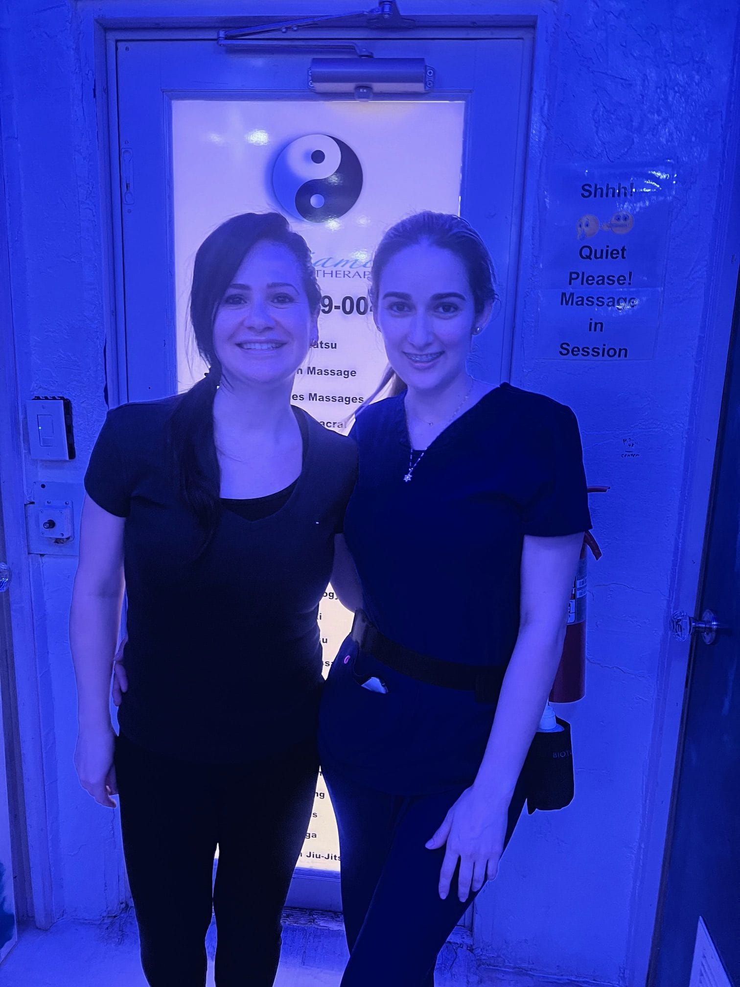 Two women stand side by side in a dimly lit room with a blue hue The door behind them features a Yin Yang symbol and a sign requesting quiet for ongoing massage sessions