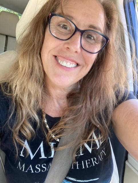 A woman with long wavy hair and glasses smiles while sitting in a car She is wearing a black T shirt with text and a seatbelt