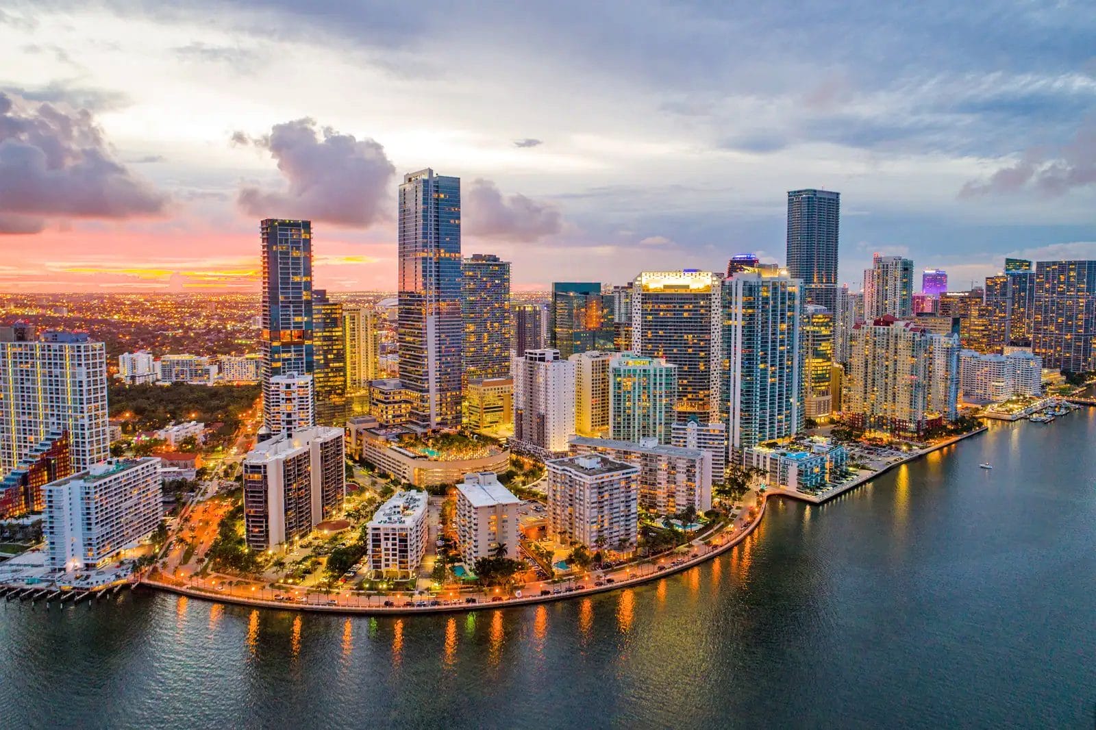 Aerial view of a modern city skyline with numerous high rise buildings lights starting to illuminate and a waterfront in the foreground at sunset