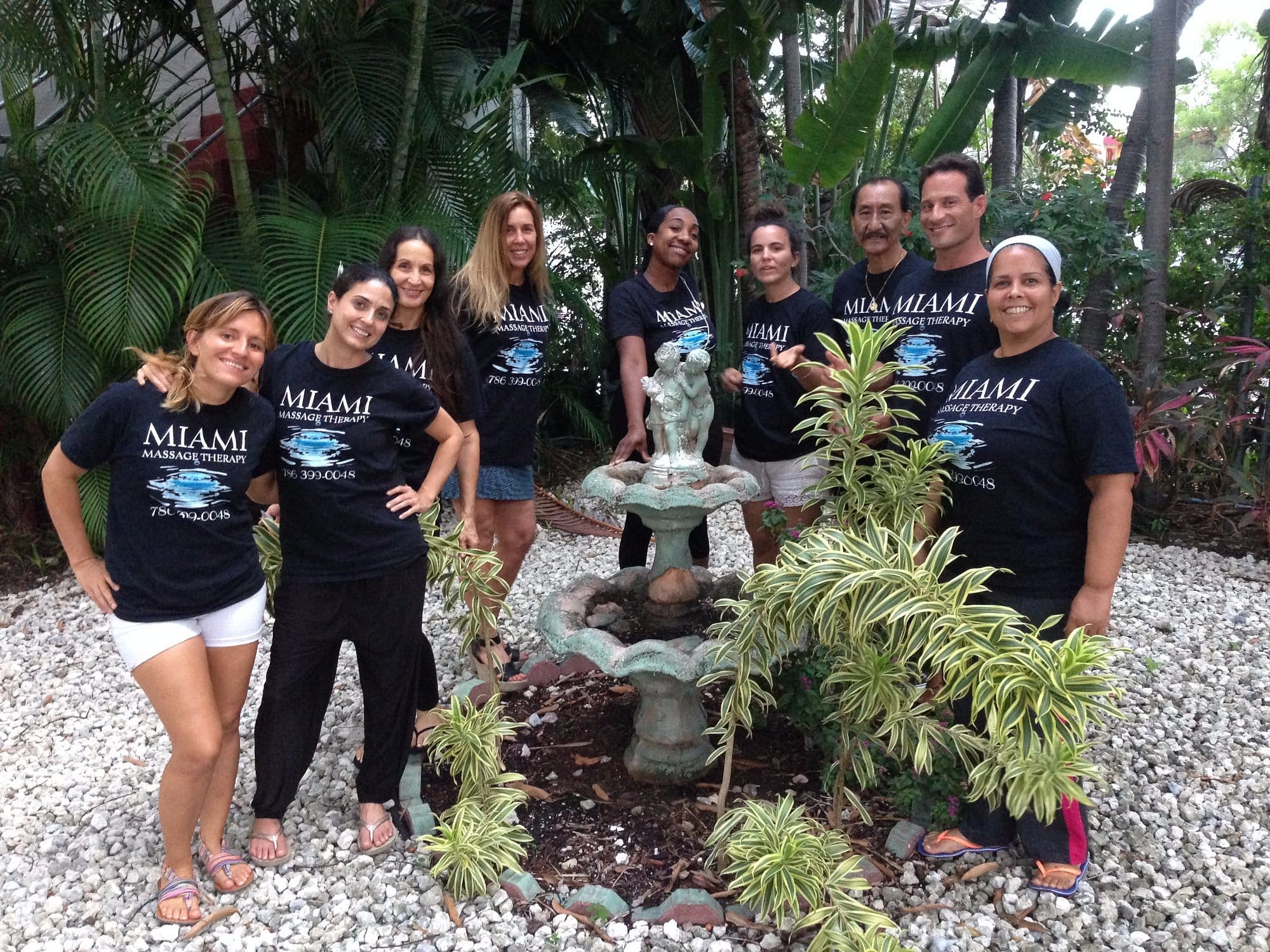 A group of nine people all wearing black Miami Massage Therapy T shirts stand around a fountain in a garden with lush greenery in the background